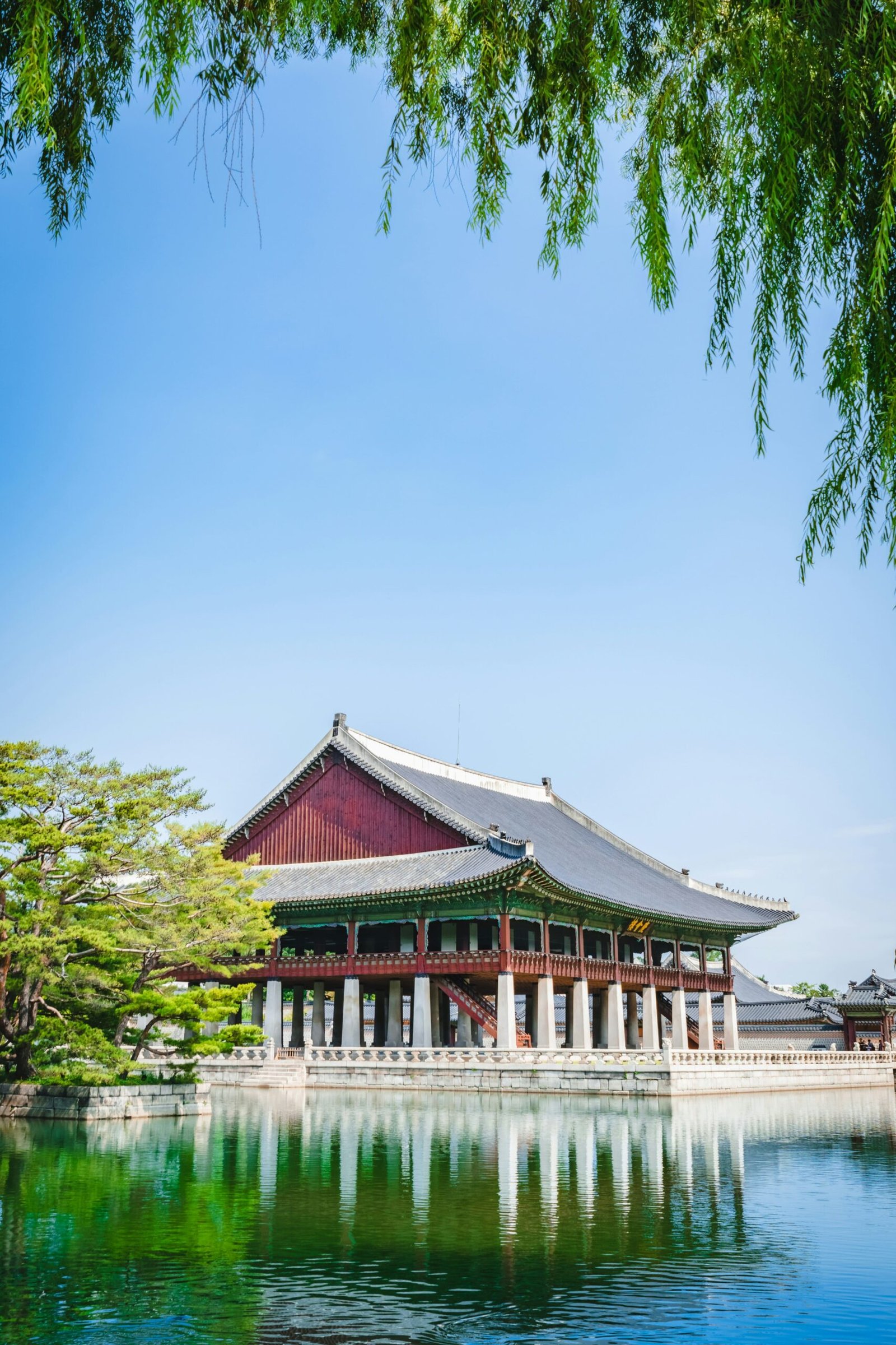 Pavillon traditionnel coréen au bord d’un étang, reflété dans l’eau sous un ciel bleu clair, encadré par des branches vertes
